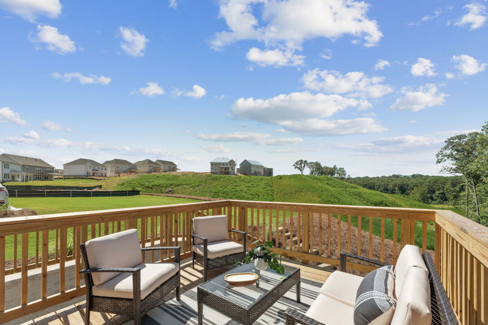 A deck with chairs and a table on it with grass and buildings in the background.